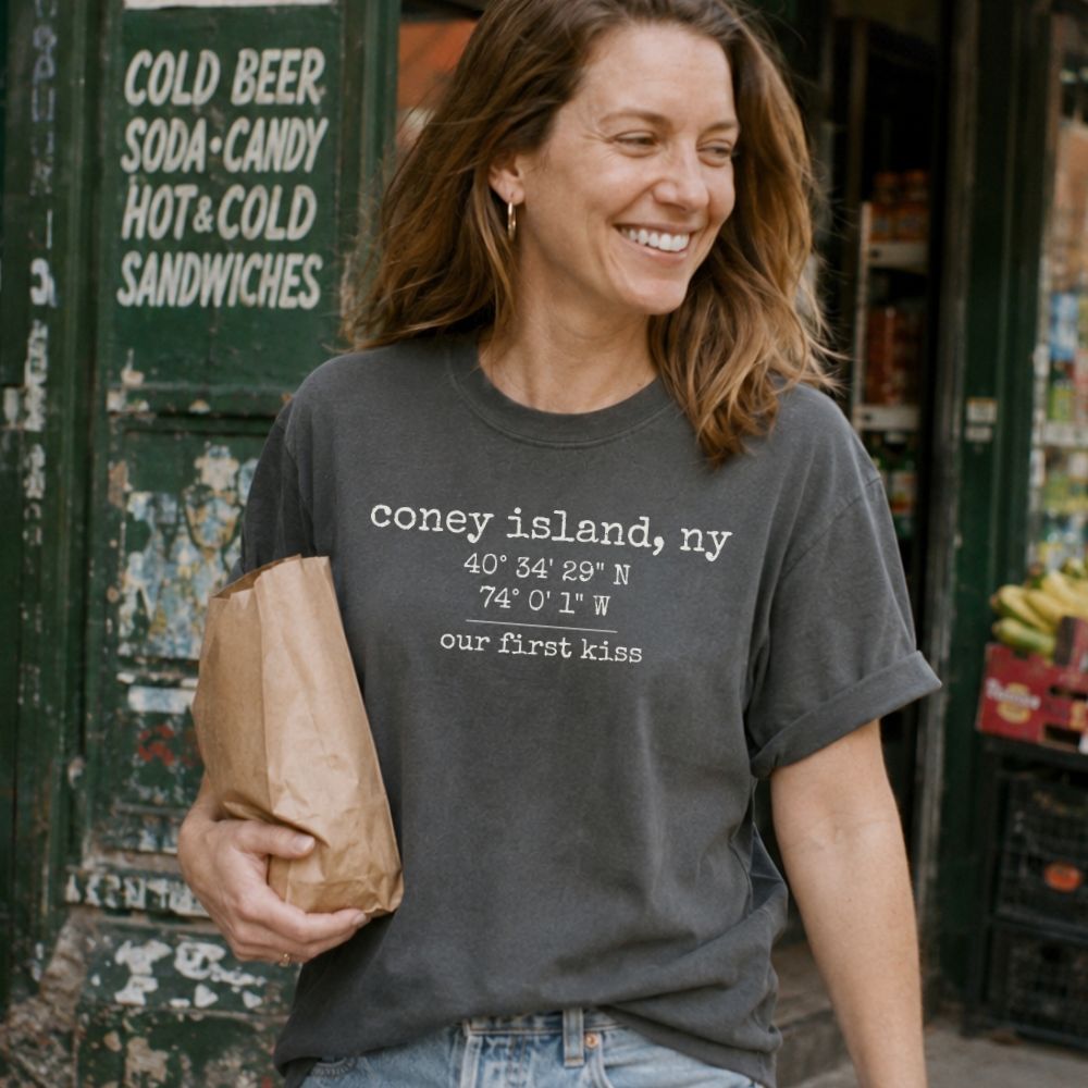 Woman wearing a Pepper colored Comfort Colors t-shirt with Coney Island coordinates, standing in front of a store.