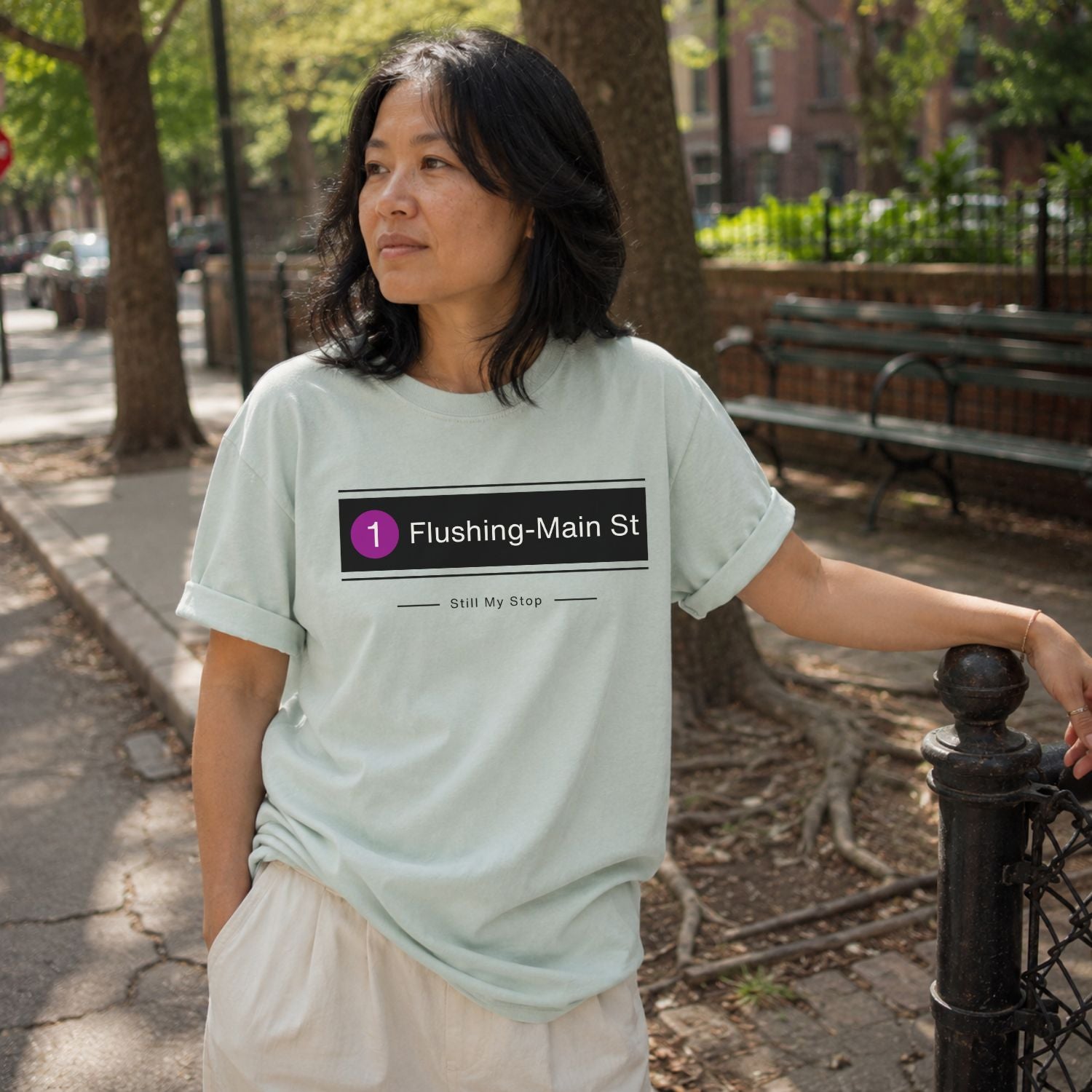 Person wearing a Bay-colored Comfort Colors t-shirt with 'Flushing-Main St' design, standing outdoors.
