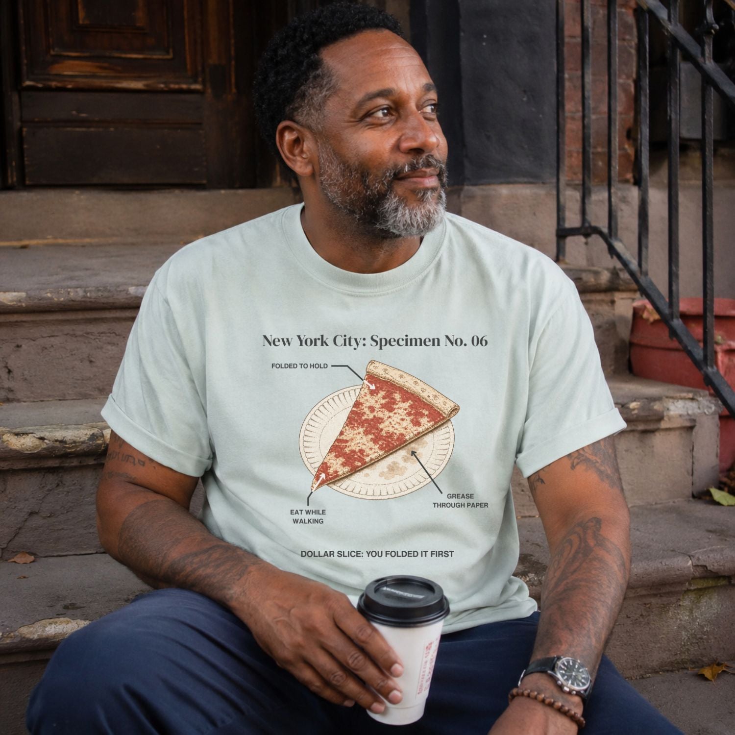 Man wearing a Bay colored Comfort Colors t-shirt featuring a vintage New York City dollar slice pizza graphic with specimen-style callouts and a folded slice on a paper plate.sitting on steps.