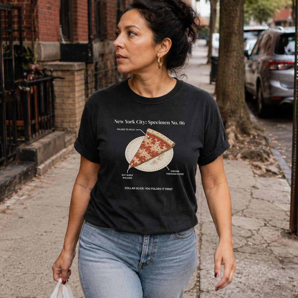 Woman walking on a sidewalk wearing a Black Comfort Colors t-shirt featuring a vintage New York City dollar slice pizza graphic with specimen-style callouts and a folded slice on a paper plate.