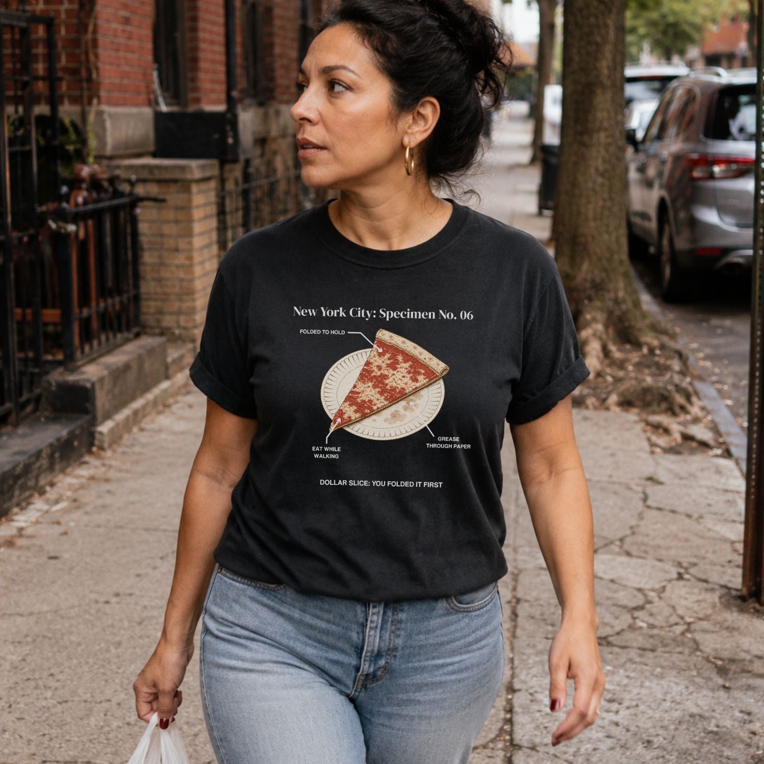 Woman walking on a sidewalk wearing a Black Comfort Colors t-shirt featuring a vintage New York City dollar slice pizza graphic with specimen-style callouts and a folded slice on a paper plate.