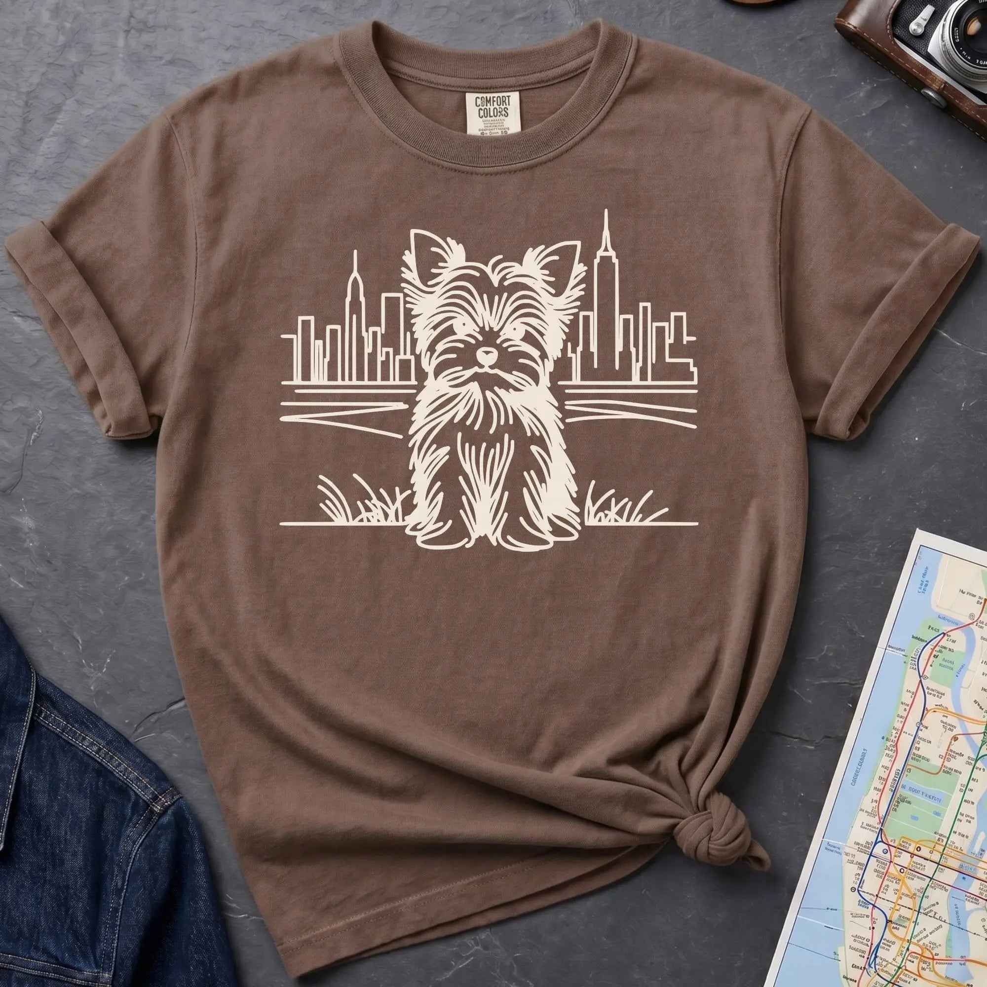 Brown Comfort Colors t-shirt featuring a minimalist Yorkshire Terrier (Yorkie) illustration in front of the New York City skyline.