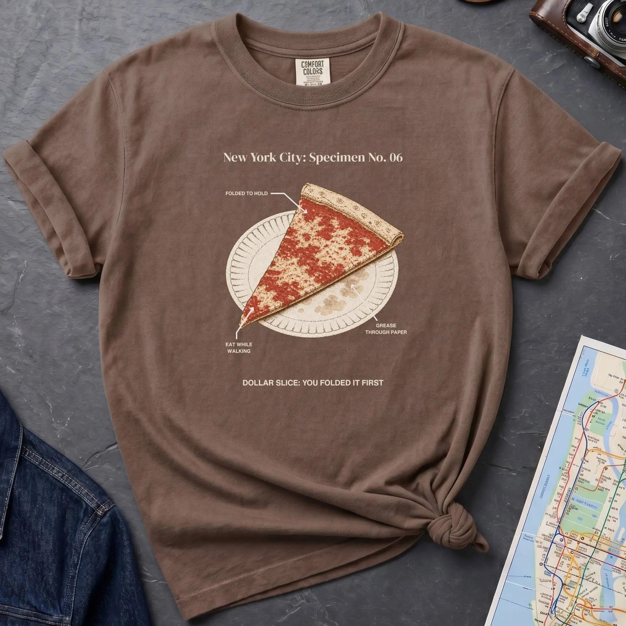 Brown Comfort Colors t-shirt featuring a vintage New York City dollar slice pizza graphic with specimen-style callouts and a folded slice on a paper plate.
