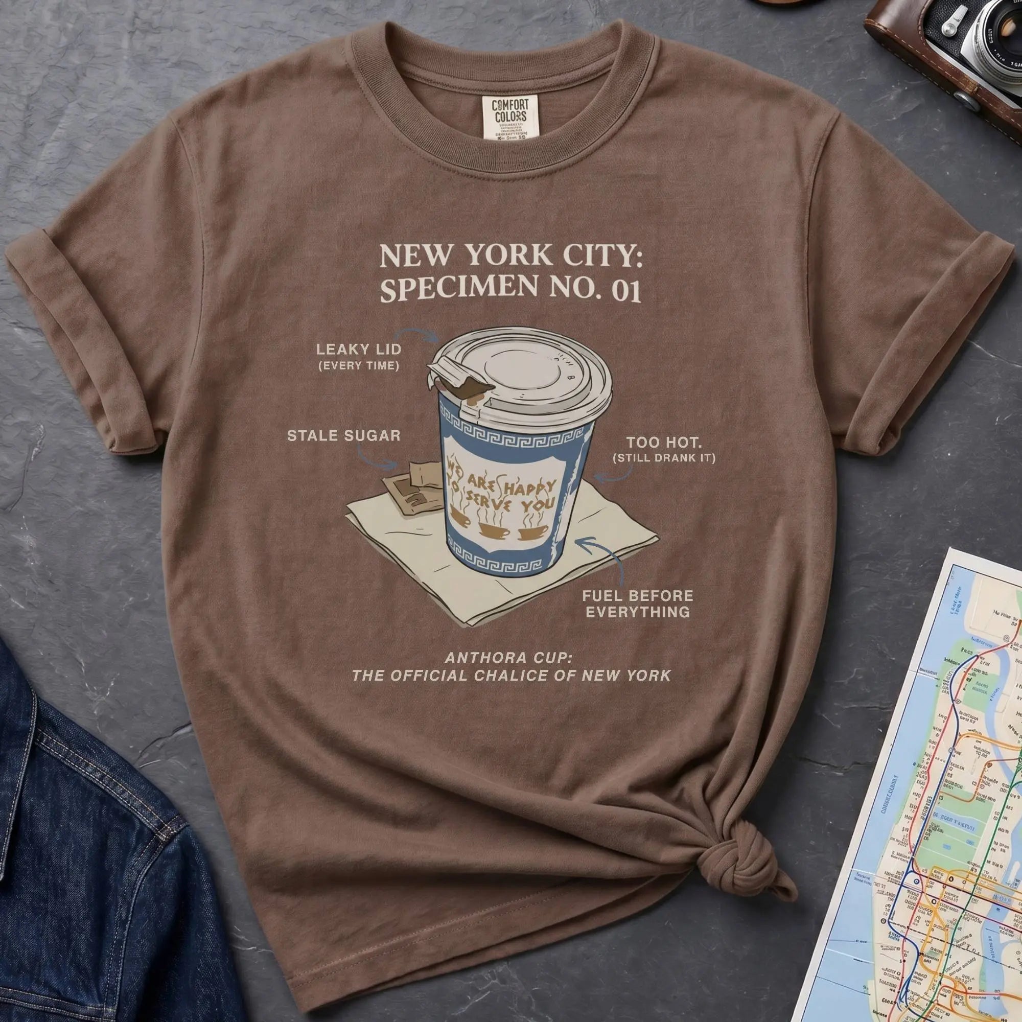 Brown t-shirt with New York City coffee cup graphic on a textured surface with map and camera.