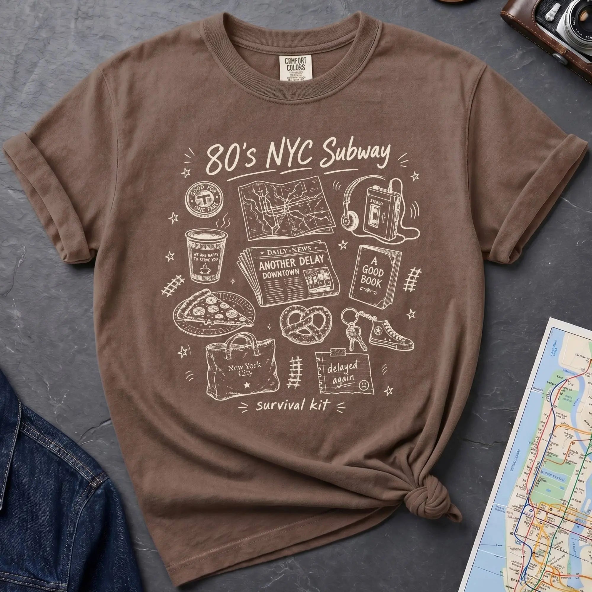 Brown t-shirt with '80s NYC Subway Survival Kit' graphic design on a dark surface with a map and denim jacket.
