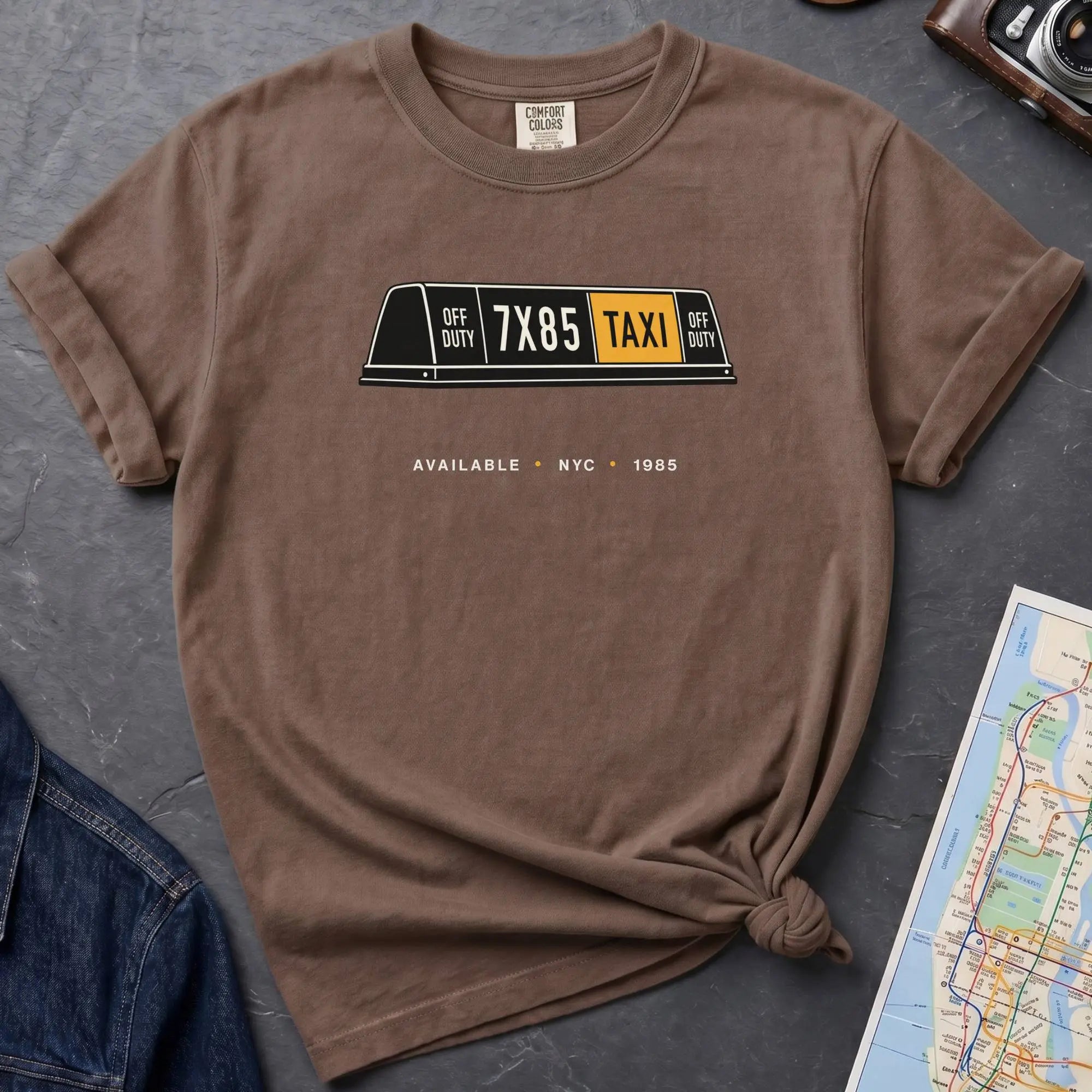 Brown Comfort Colors t-shirt featuring a vintage New York City taxi roof sign graphic with “Available • NYC • 1985” text.