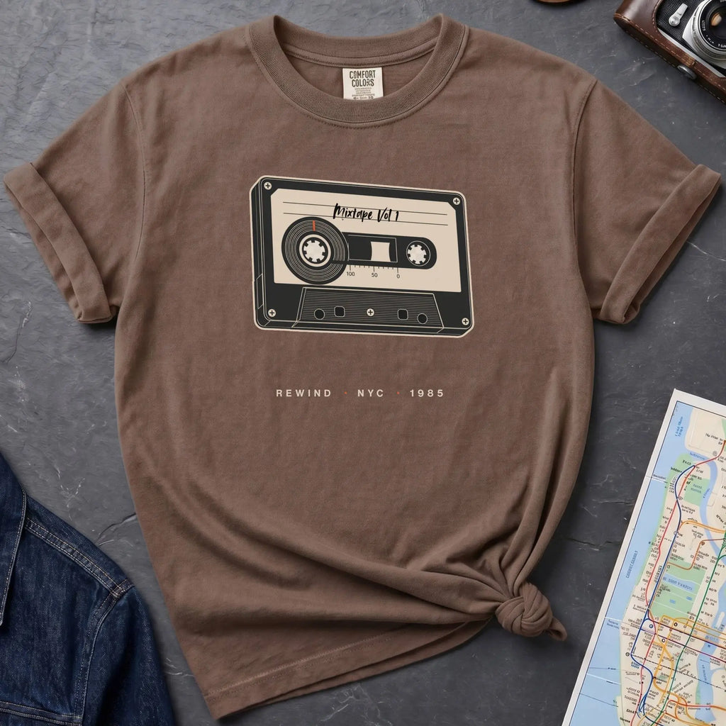 Brown t-shirt with cassette tape graphic and text on a gray surface with denim jacket and map.