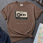 Brown t-shirt with cassette tape graphic and text on a gray surface with denim jacket and map.