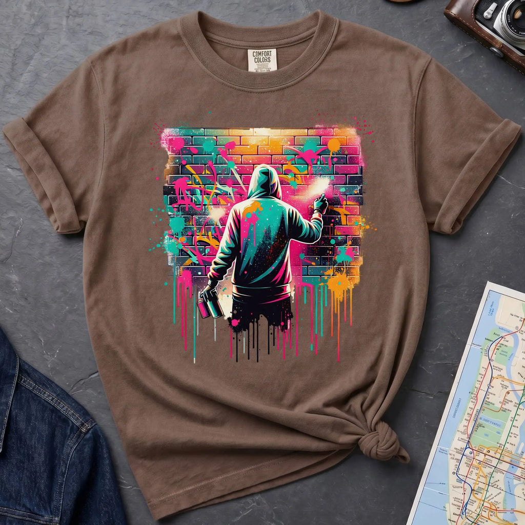 Brown t-shirt with a graphic of a person spray painting a colorful mural on a brick wall.