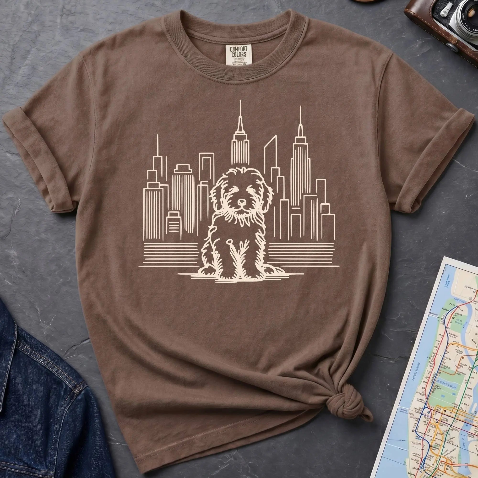 Brown t-shirt with a Golden Doodle and cityscape design on a dark surface with a map and camera.