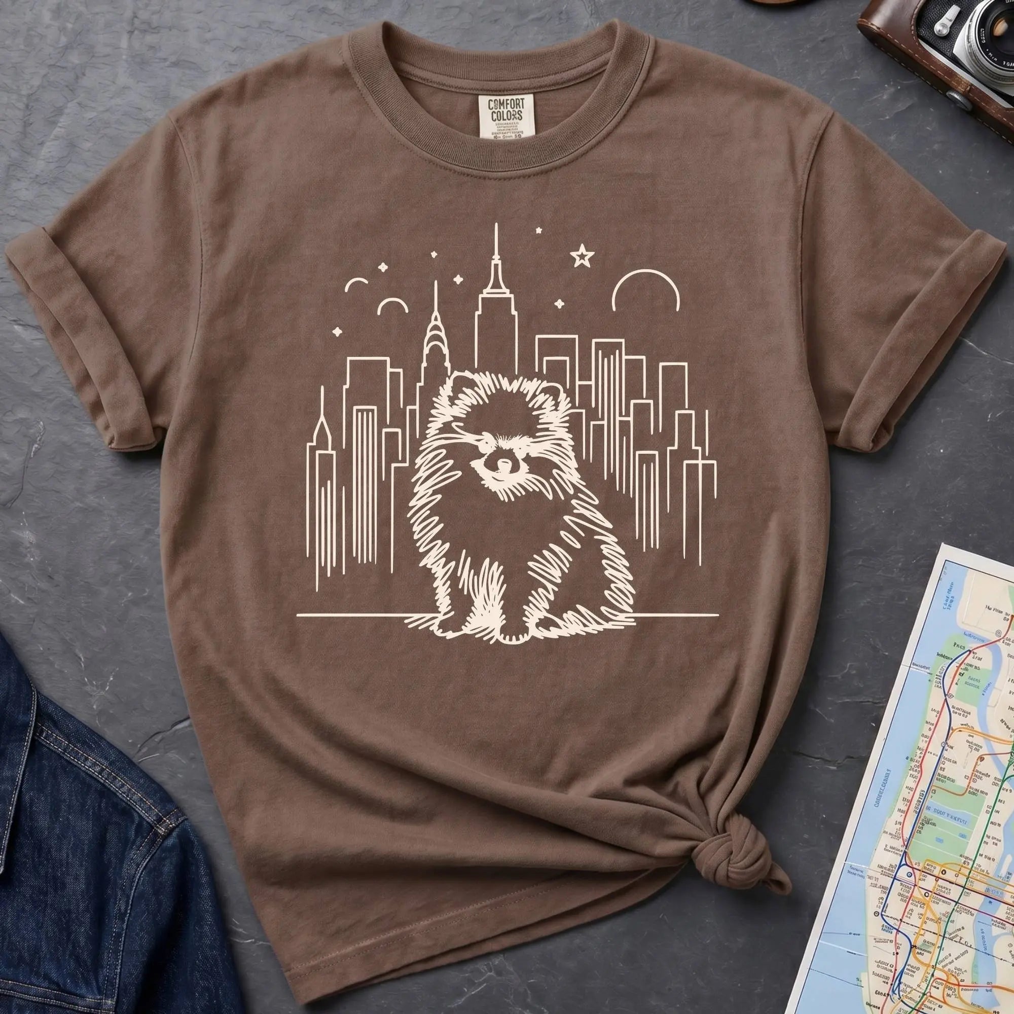 Brown Comfort Colors t-shirt featuring a fluffy small dog illustration in front of the New York City skyline.