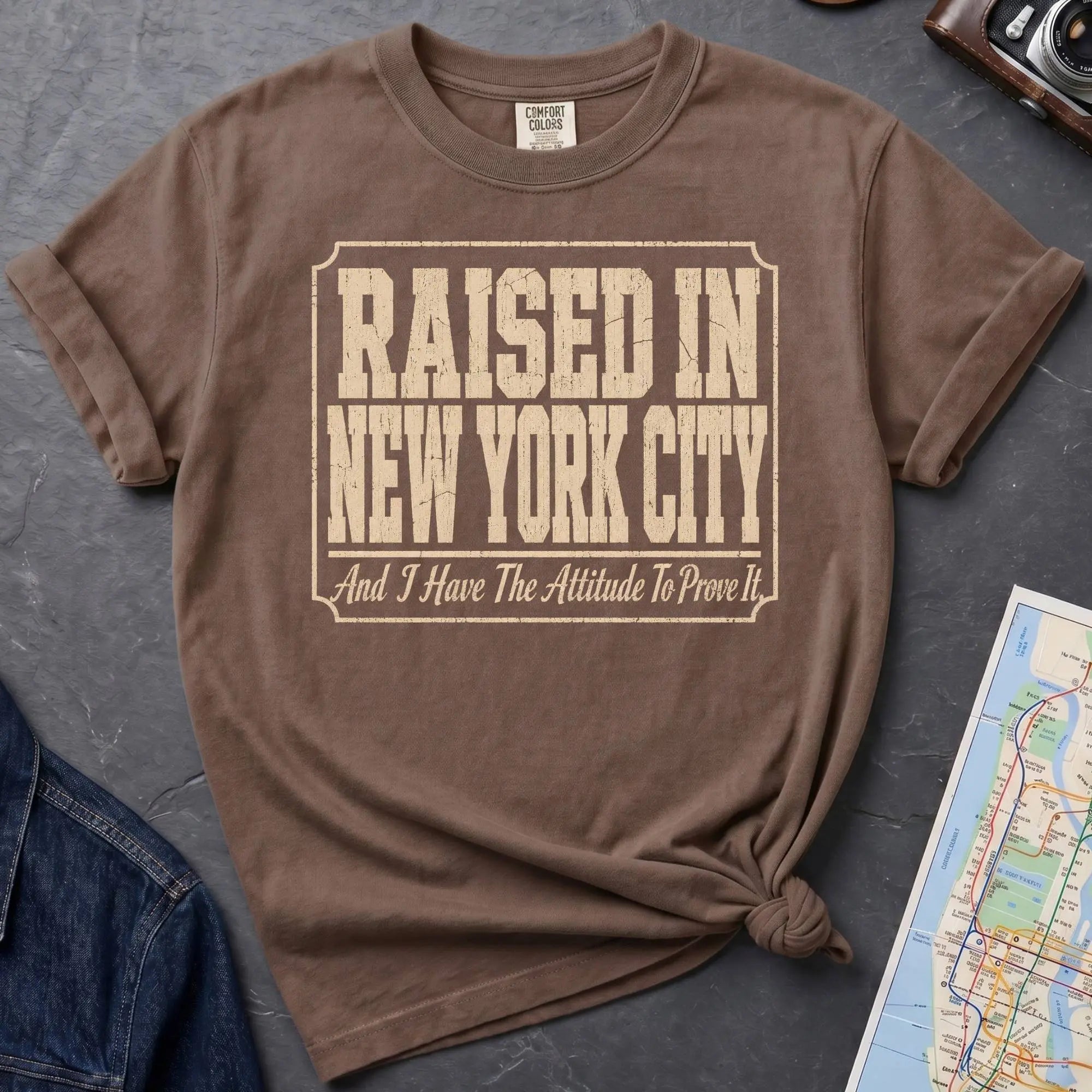 Brown Comfort Colors t-shirt featuring distressed vintage text that reads “Raised in New York City, And I Have the Attitude to Prove It.