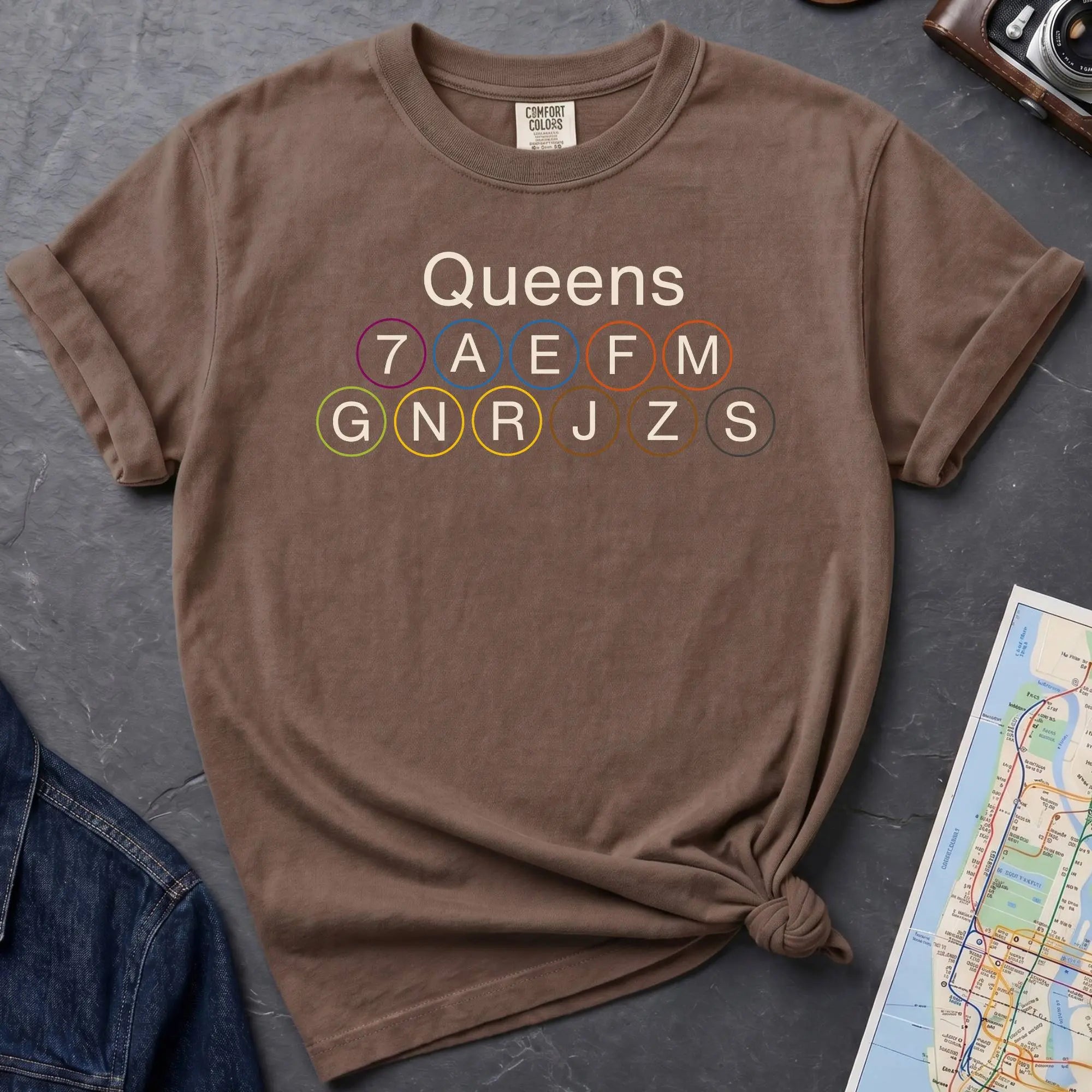 Brown Comfort Colors t-shirt featuring a minimalist Queens subway line graphic with Queens text and classic NYC train icons.