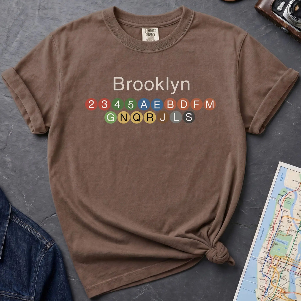 Brown t-shirt with 'Brooklyn' and subway line numbers on a dark surface with a map and camera.