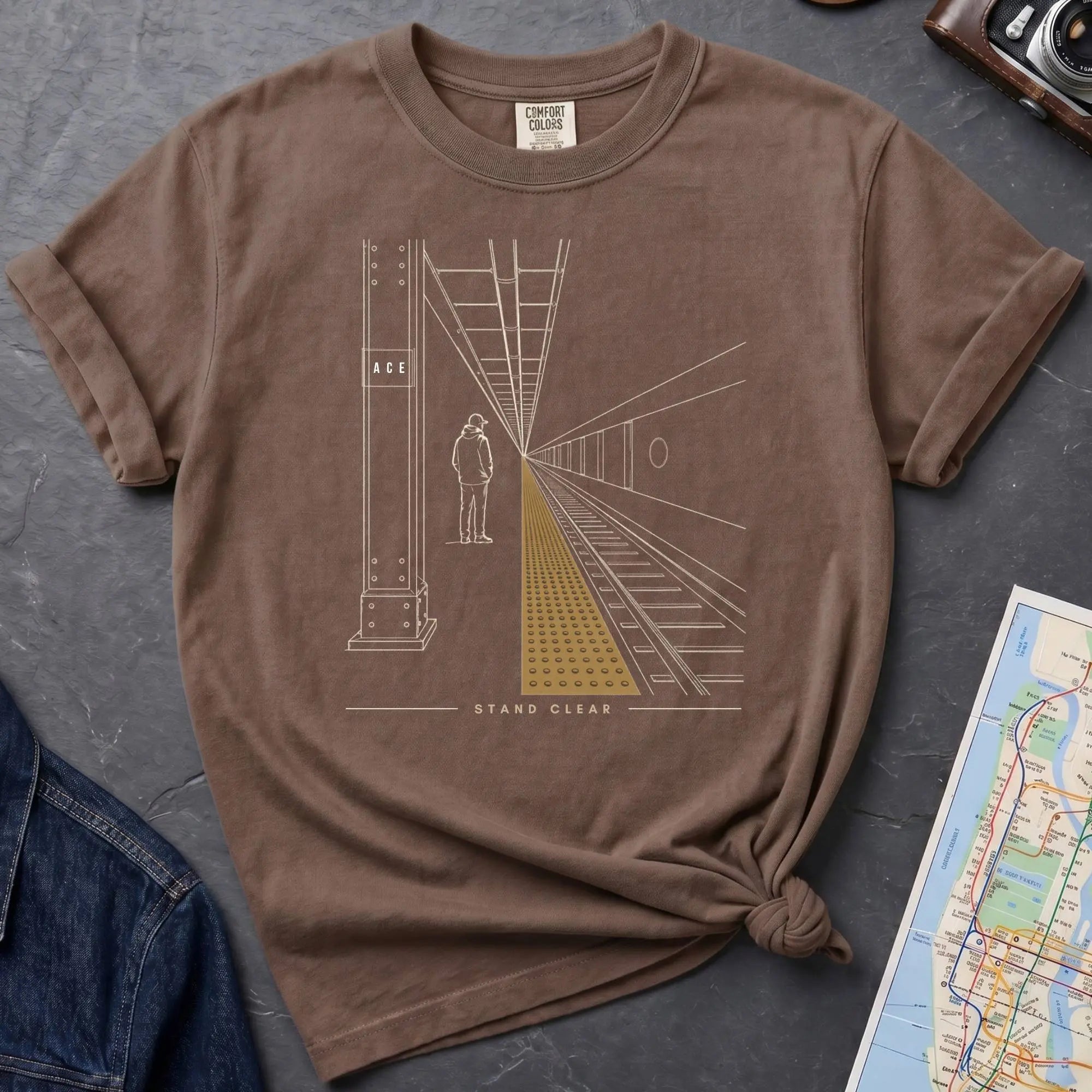 Brown Comfort Colors t-shirt featuring a minimalist NYC subway platform graphic with a lone figure, train tracks, and “Stand Clear” text.