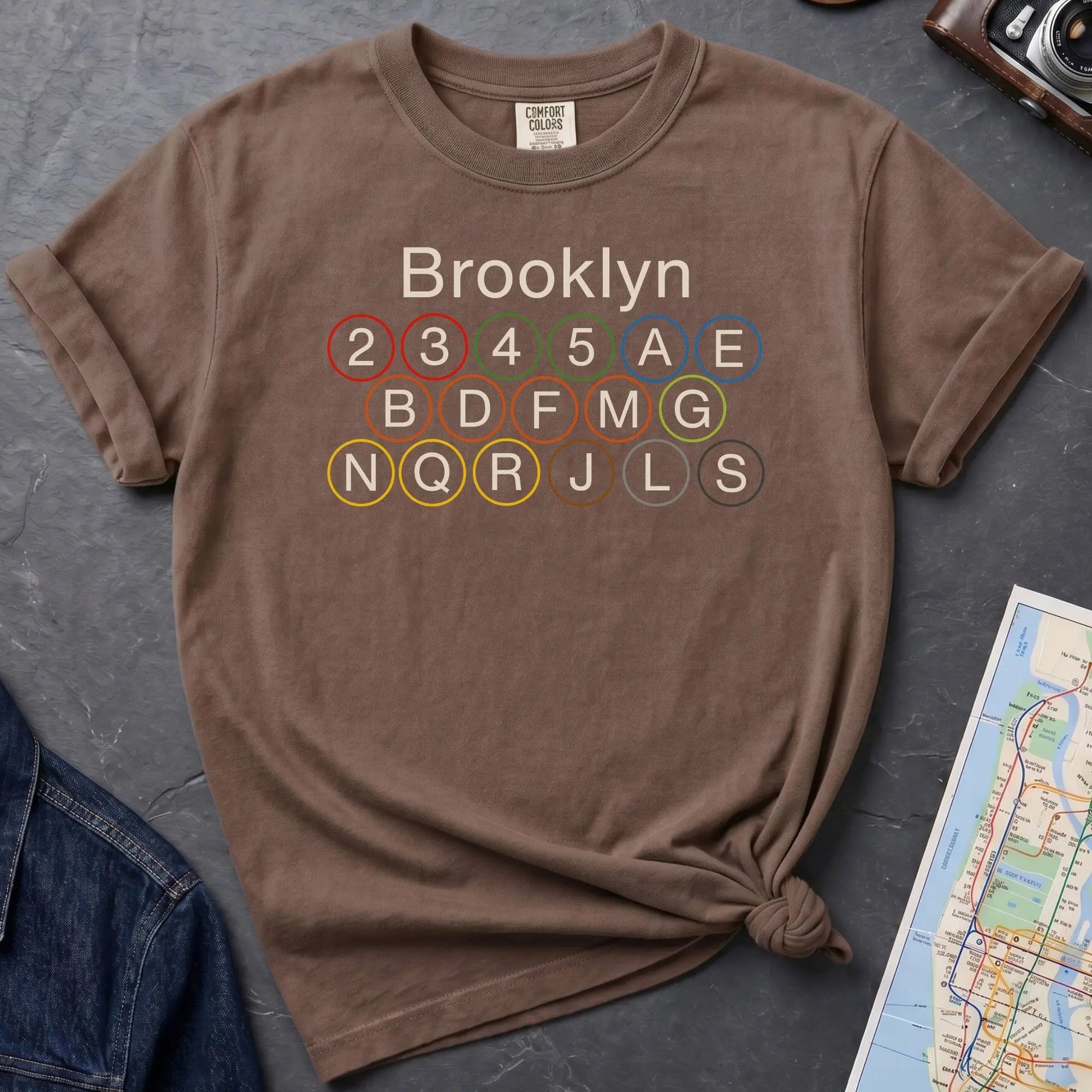 Brown Comfort Colors t-shirt featuring the word “Brooklyn” above colorful NYC subway line circles representing the subway routes that run through Brooklyn.