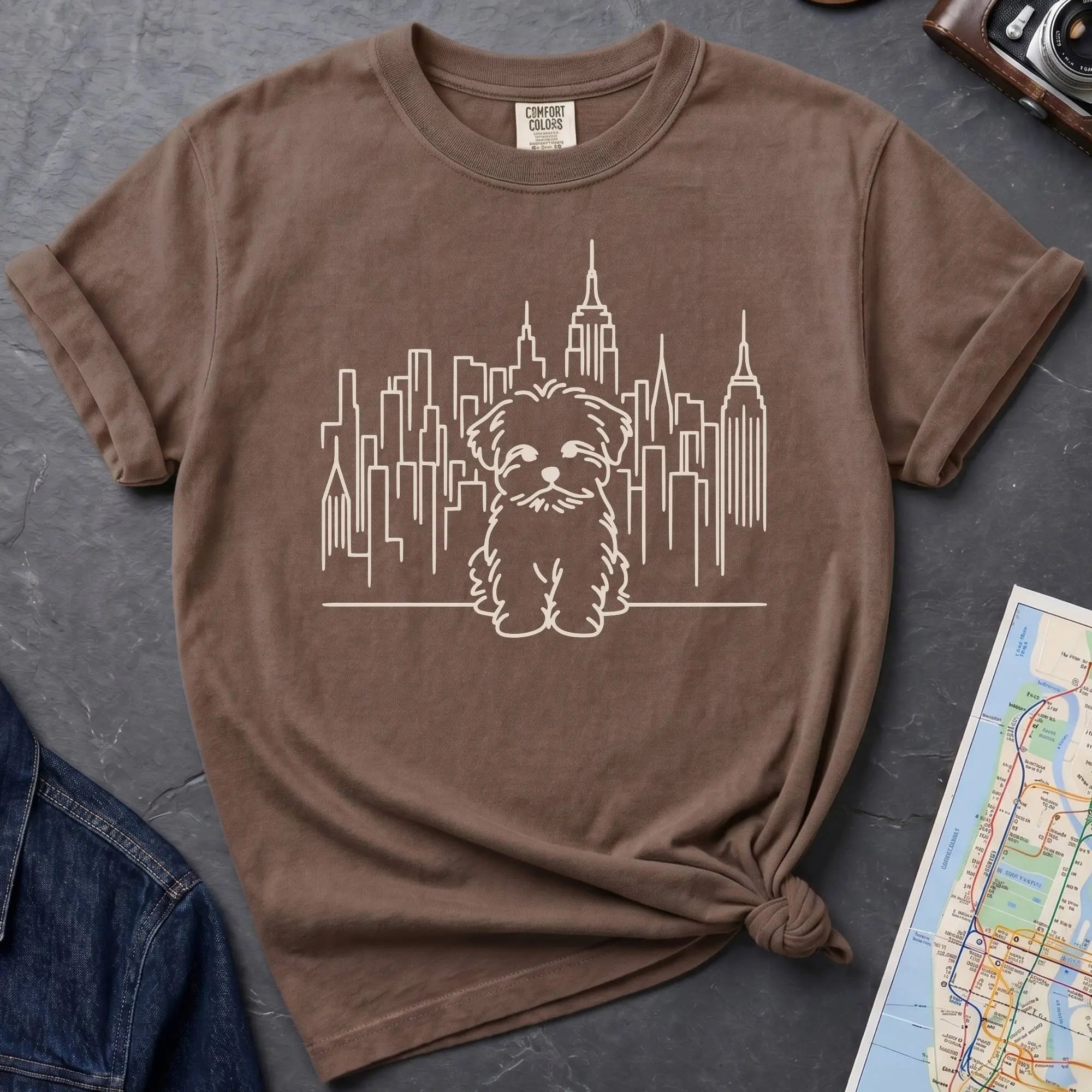 Brown t-shirt with a Maltese dog and cityscape design on a gray surface with a map and camera.