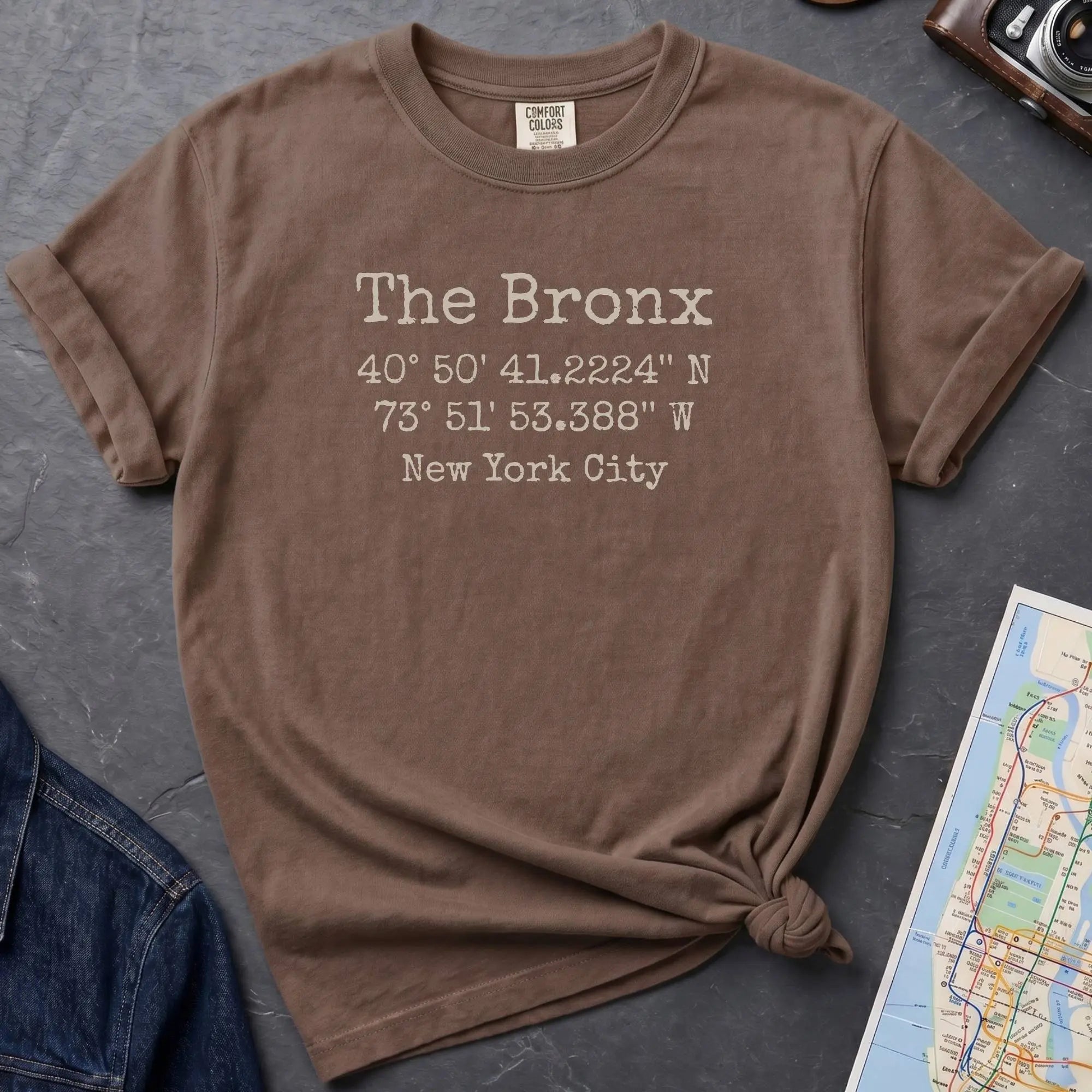 Brown Comfort Colors t-shirt featuring The Bronx, NY coordinates and New York City text in a minimalist typewriter-style design.