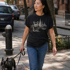 Woman walking a French Bulldog on a leash wearing a Black Comfort Colors t-shirt with a French Bulldog and cityscape design in an urban setting