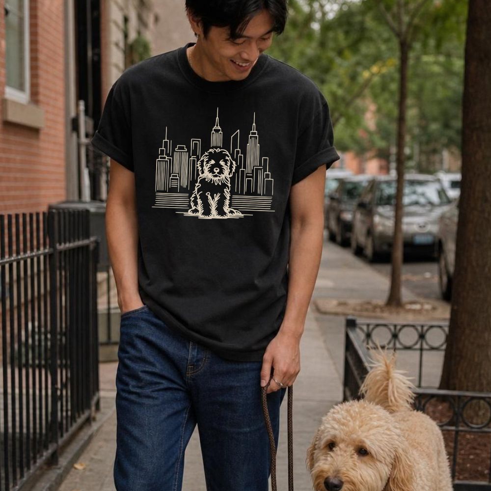 Man wearing a black Comfort Colors t-shirt with a Golden Doodle and cityscape design, walking down a sidewalk with his dog on a leash.