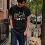 Man wearing a black Comfort Colors t-shirt with a Golden Doodle and cityscape design, walking down a sidewalk with his dog on a leash.