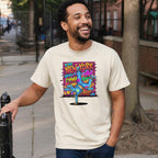 Man wearing an Ivory Comfort Colors t-shirt with a colorful graphic design featuring a breakdancer and text on a city street.