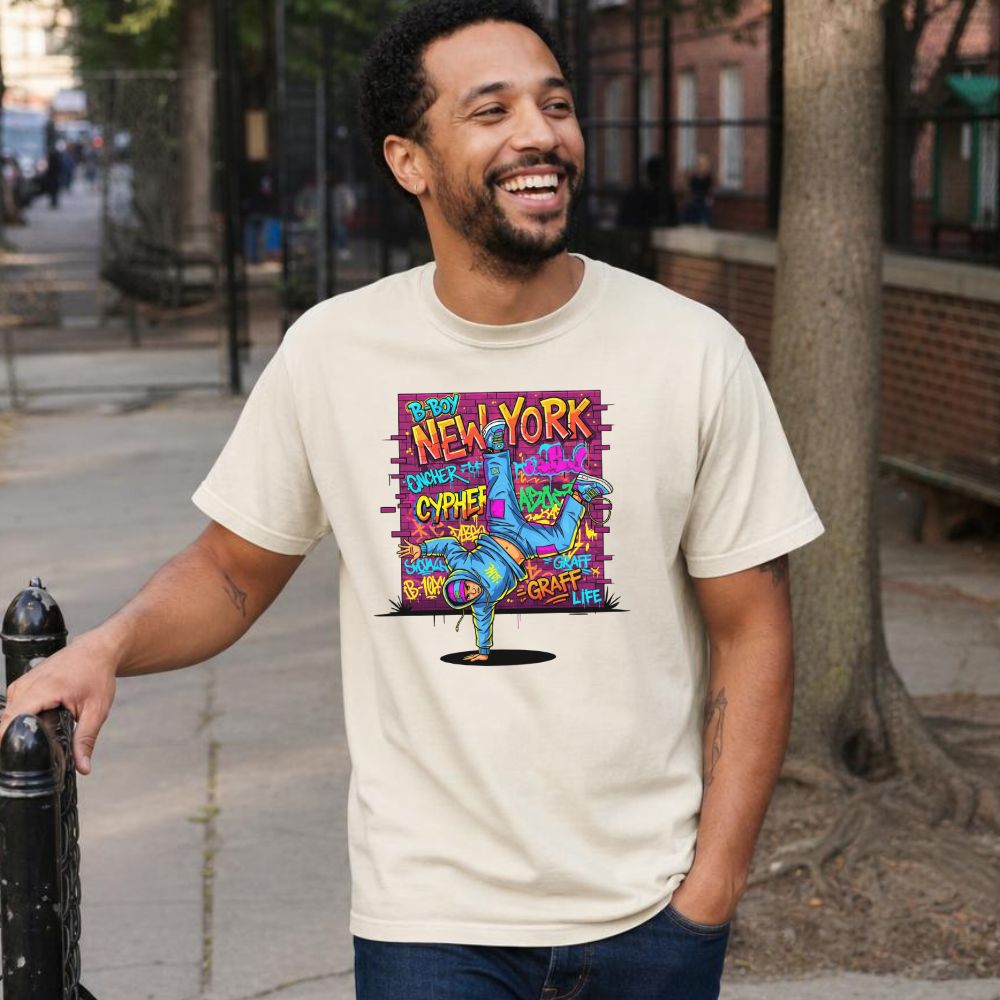 Man wearing an Ivory Comfort Colors t-shirt with a colorful graphic design featuring a breakdancer and text on a city street.