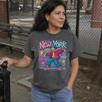 Woman wearing a pepper colored Comfort Colors t-shirt featuring a colorful NYC graffiti-style New York graphic with a young street artist holding a spray paint can design on a gray surface with denim jacket and map while standing outdoors.