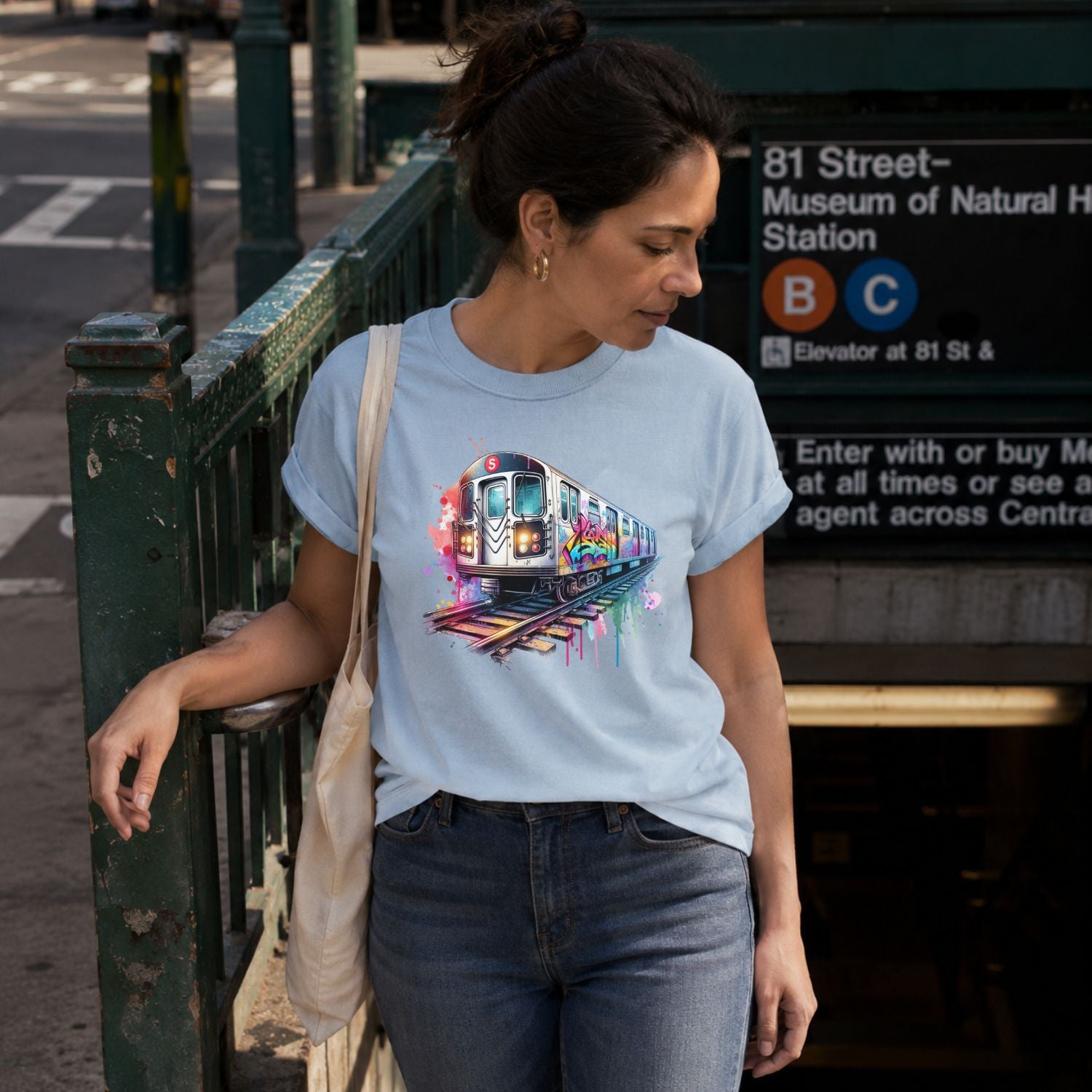Woman wearing a Light blue Comfort Colors t-shirt featuring a colorful NYC subway train graphic with graffiti-style paint details and bright street art accents walking out from a subway station.