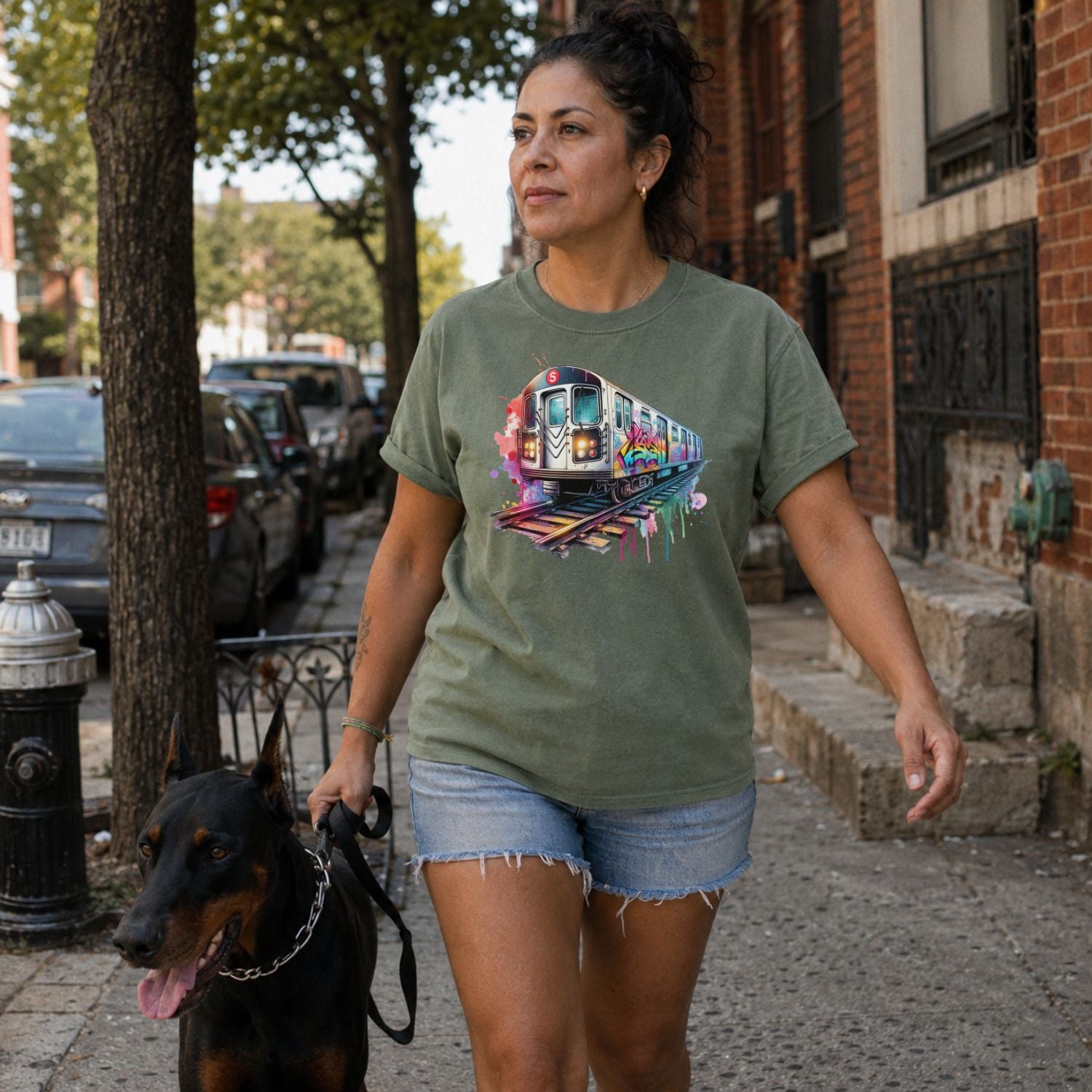 Woman walking a dog on a city street wearing a Moss green Comfort Colors t-shirt featuring a colorful NYC subway train graphic with graffiti-style paint details and bright street art accents.