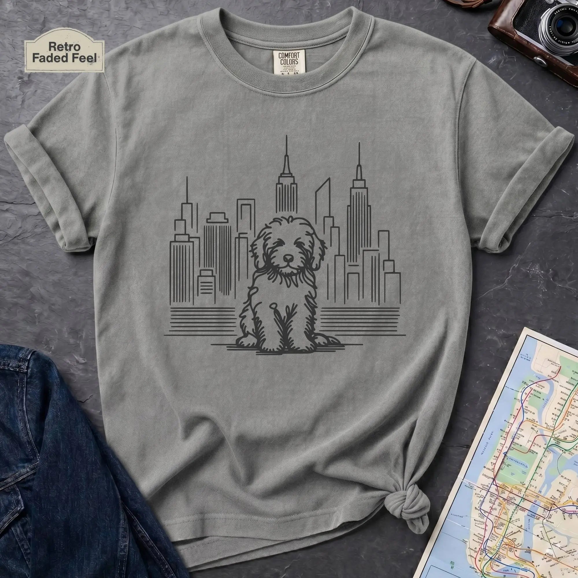 Granite colored t-shirt with a Golden Doodle dog and cityscape design on a gray surface with a map and camera.