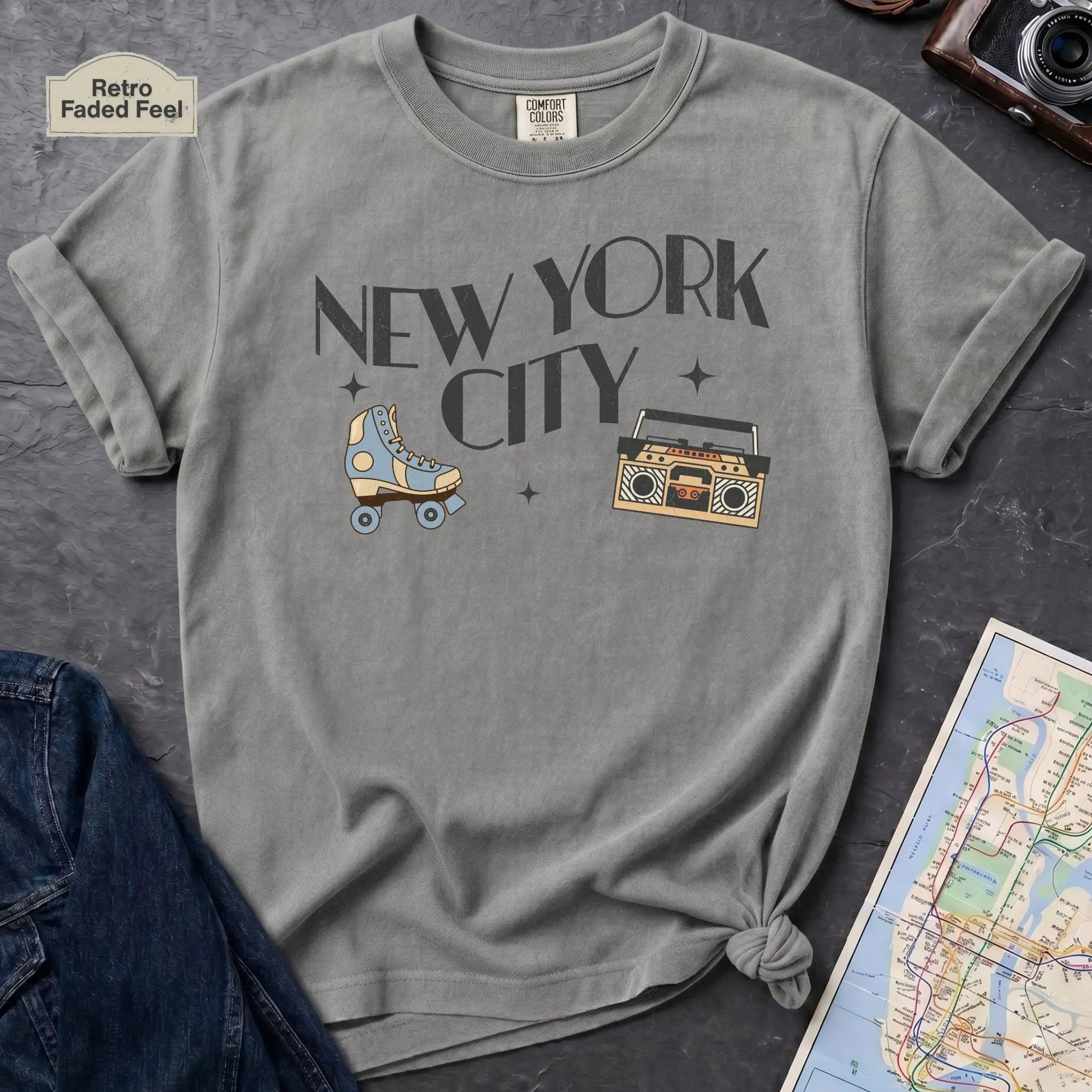 Granite colored t-shirt with 'New York City' text and a roller skate and boom box on a dark surface with a map and camera.