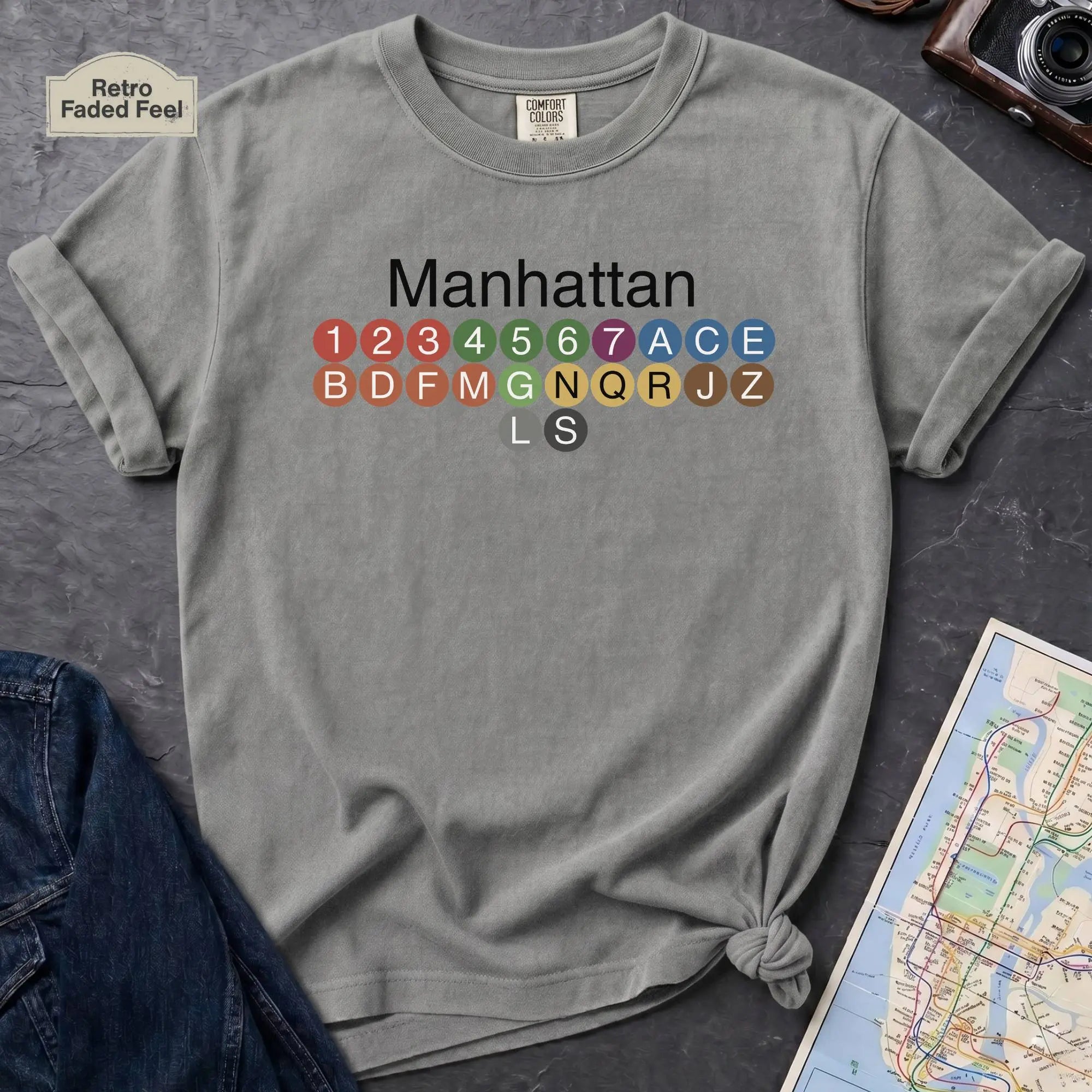 Granite colored Comfort Colors t-shirt featuring a minimalist Manhattan subway line graphic with colorful NYC train icons.