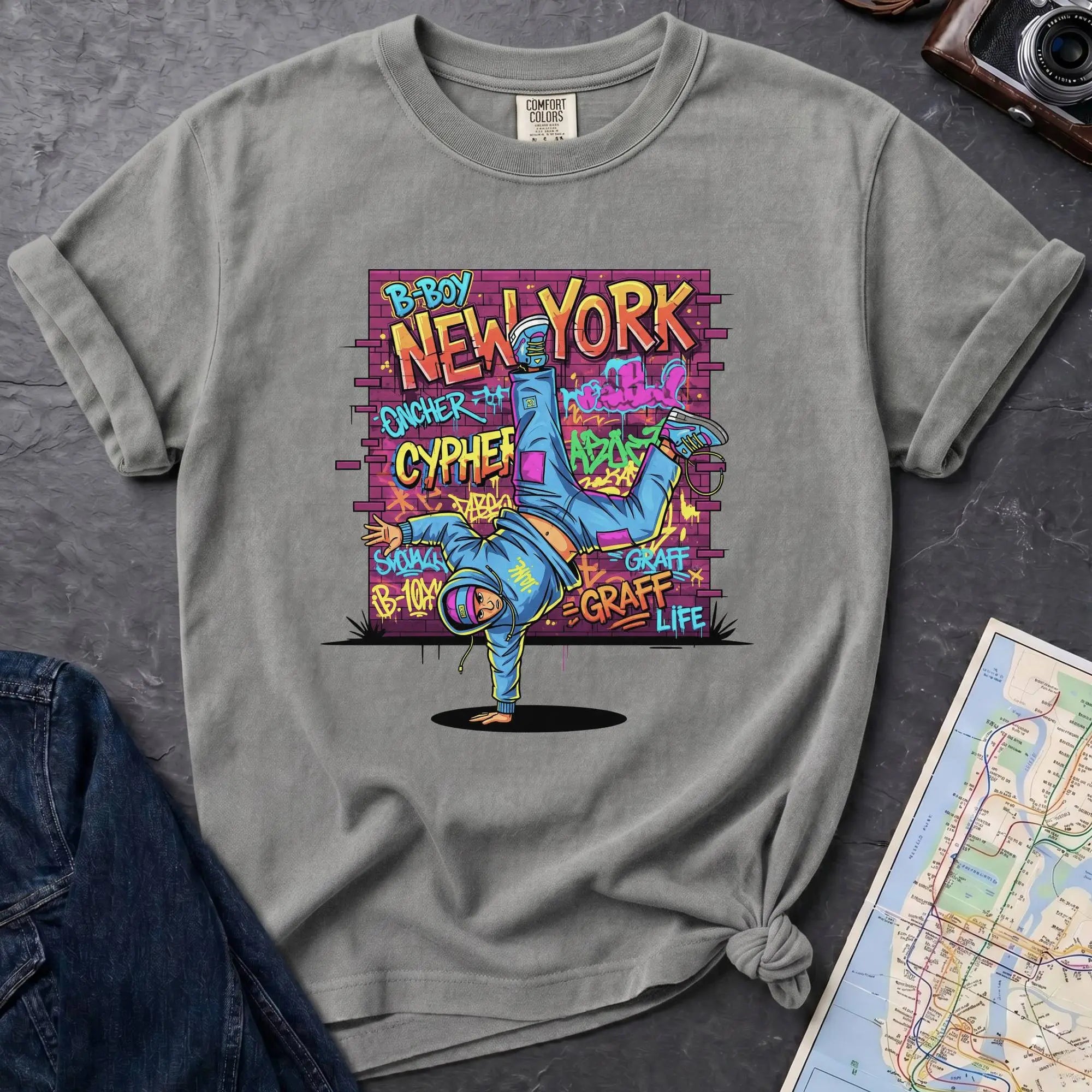 Granite colored t-shirt with a colorful graphic design featuring a breakdancer and text on a dark surface with a denim jacket and a map.