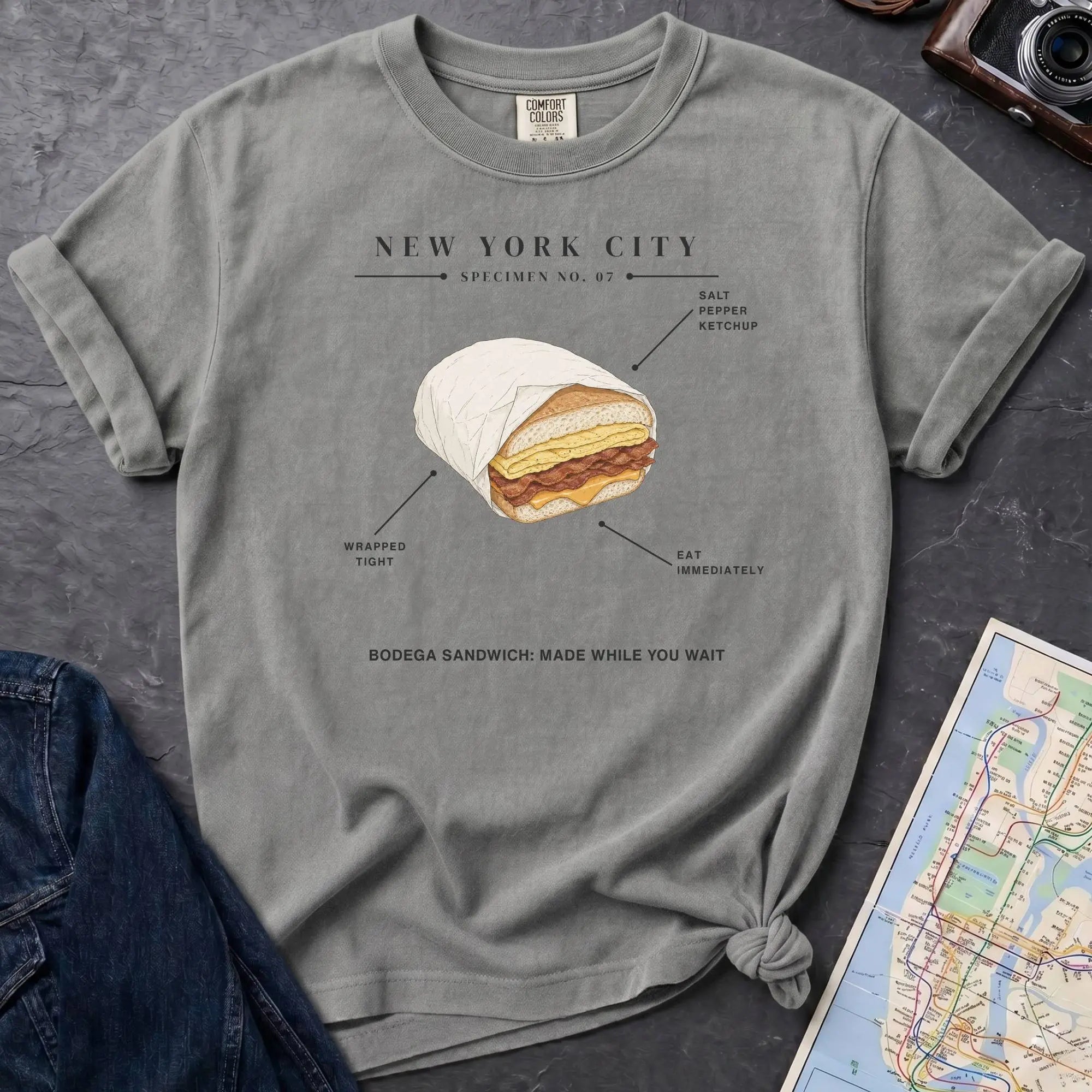Granite colored Comfort Colors t-shirt featuring a New York City bodega sandwich graphic with specimen-style callouts and “Bodega Sandwich: Made While You Wait” text.