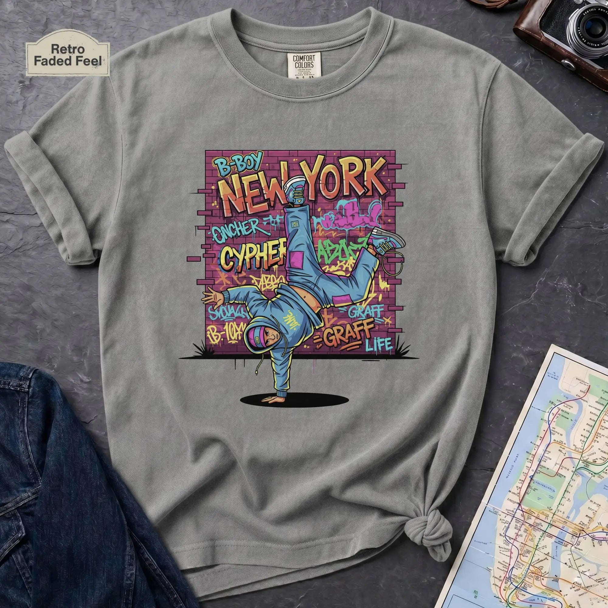 Granite colored t-shirt with a colorful graphic design featuring a breakdancer and text on a dark surface with a denim jacket and a map.