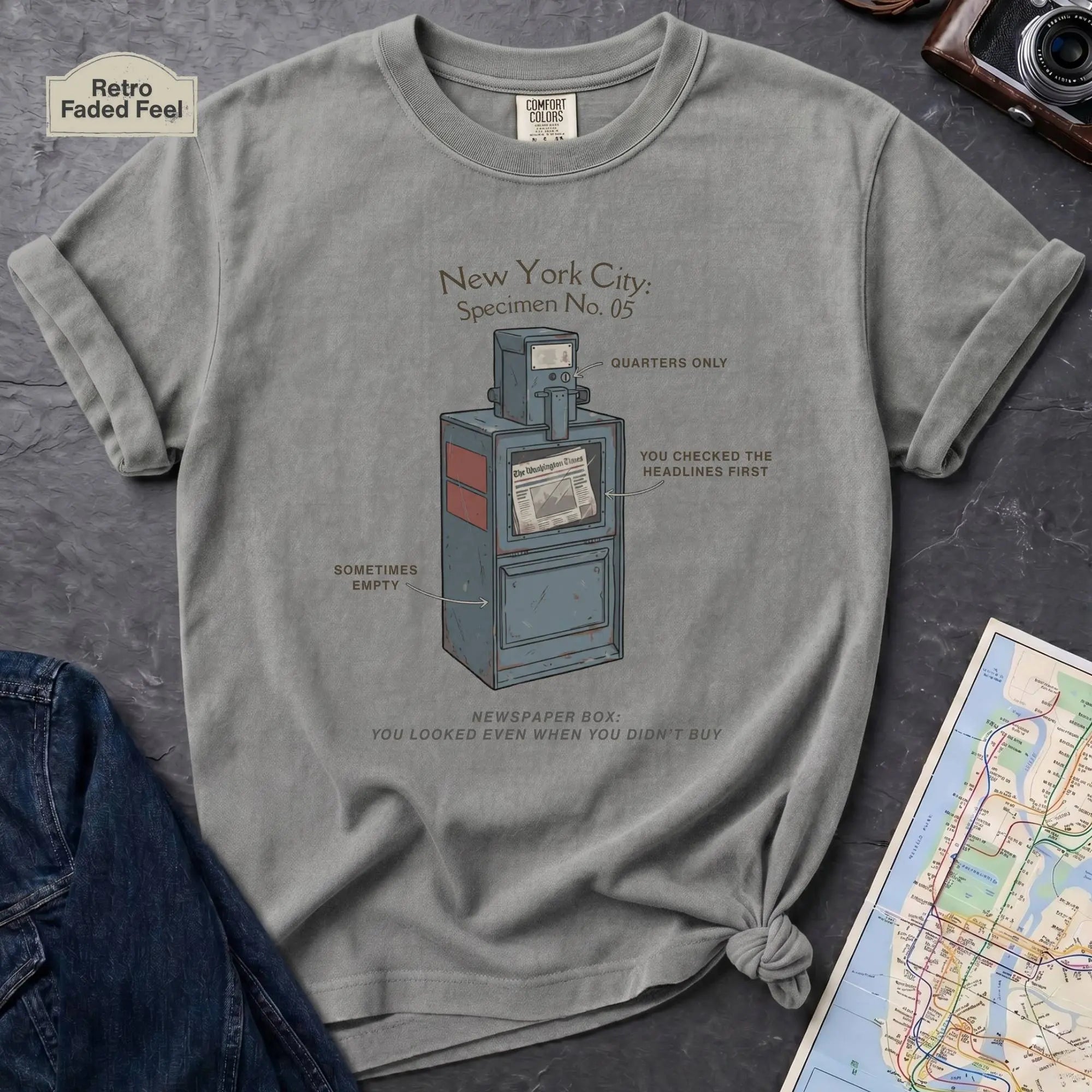 Granite colored t-shirt with a graphic design of a vintage newspaper box and text on a gray surface with a map and camera.