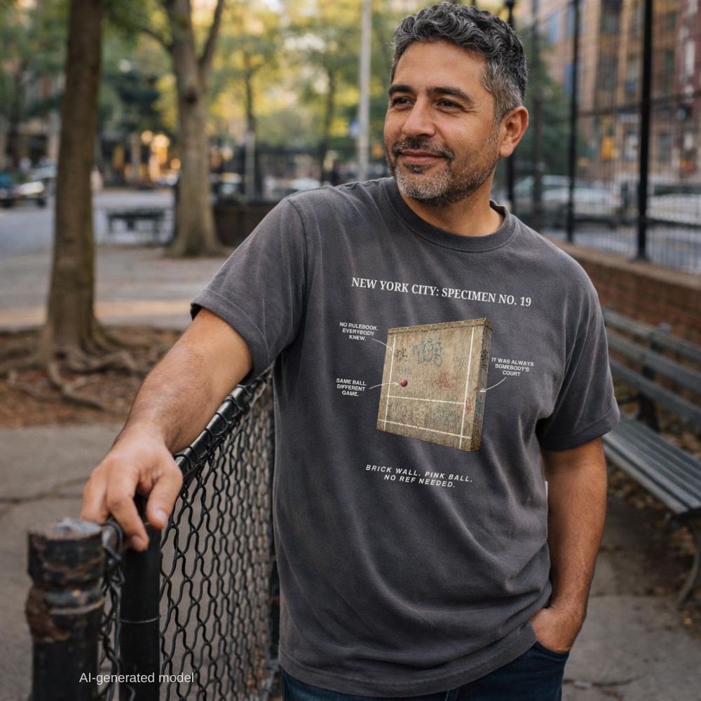 Man wearing a pepper-colored Comfort Colors t-shirt featuring a vintage scientific specimen illustration of a graffiti covered NYC handball court wall with a pink Spaldeen ball and callout arrows labeled No Rulebook Everybody Knew, Same Ball Different Game, It Was Always Somebody's Court, with tagline: Brick Wall, Pink Ball, No Ref Needed, while leaning on a park fence.