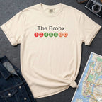 Beige t-shirt with 'The Bronx' text and NYC subway lines on a dark surface with a denim jacket, camera and a map.