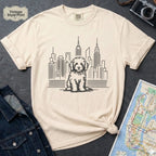 Ivory t-shirt with a Golden Doodle dog and cityscape design on a gray surface with a map and camera.