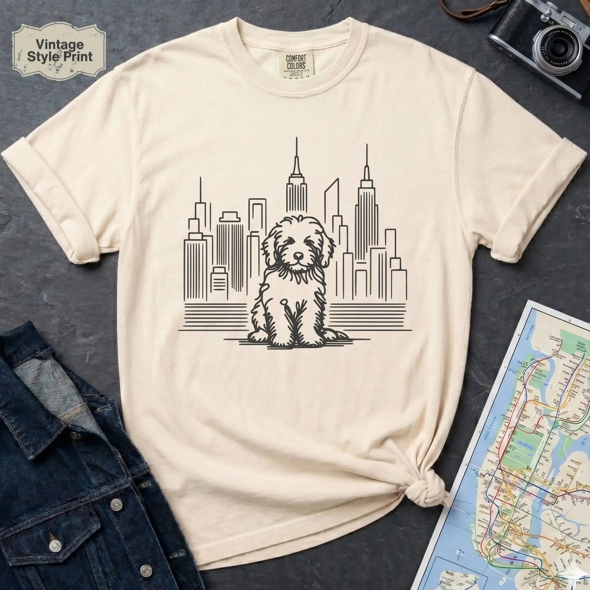 Ivory t-shirt with a Golden Doodle dog and cityscape design on a gray surface with a map and camera.