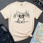 Ivory t-shirt with a Shih Tzu dog and cityscape design on a dark surface with a map and camera.