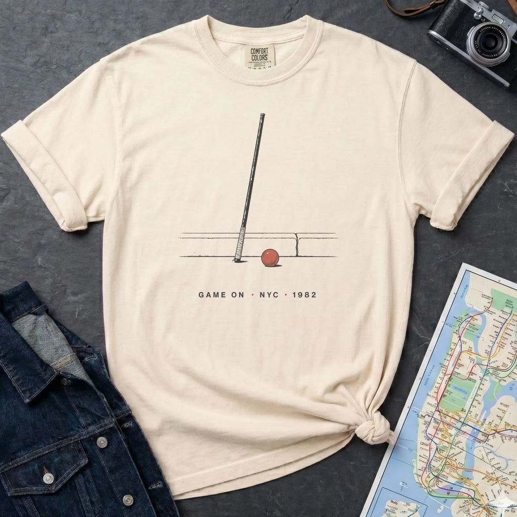 Ivory Comfort Colors t-shirt featuring a minimalist stickball bat and ball graphic with “Game On • NYC • 1982” text.