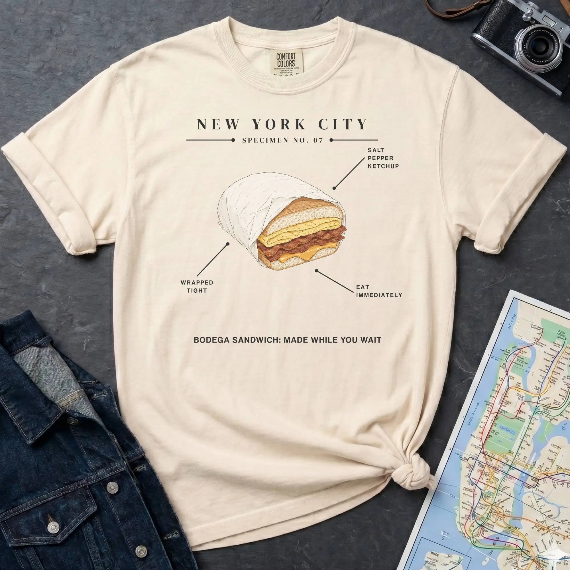 Ivory Comfort Colors t-shirt featuring a New York City bodega sandwich graphic with specimen-style callouts and “Bodega Sandwich: Made While You Wait” text.