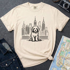 Ivory t-shirt with a Golden Doodle dog and cityscape design on a gray surface with a map and camera.