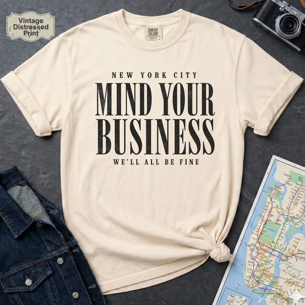 Ivory Comfort Colors t-shirt featuring bold New York City “Mind Your Business, We’ll All Be Fine” text in a vintage black typography design.