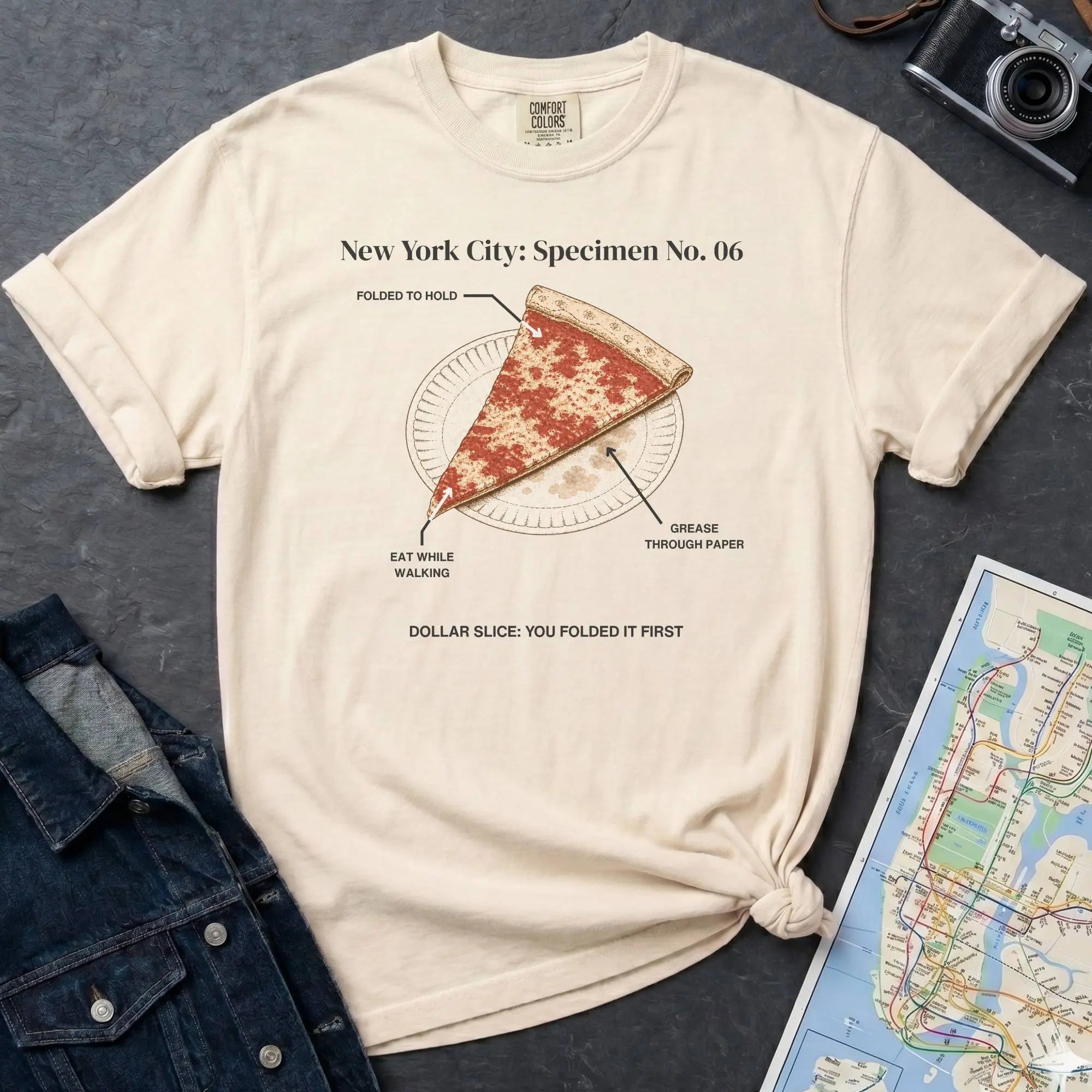 Ivory Comfort Colors t-shirt featuring a vintage New York City dollar slice pizza graphic with specimen-style callouts and a folded slice on a paper plate.