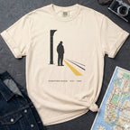 Ivory t-shirt with a graphic design of a person on a train platform, placed on a dark surface with a map and denim jacket.