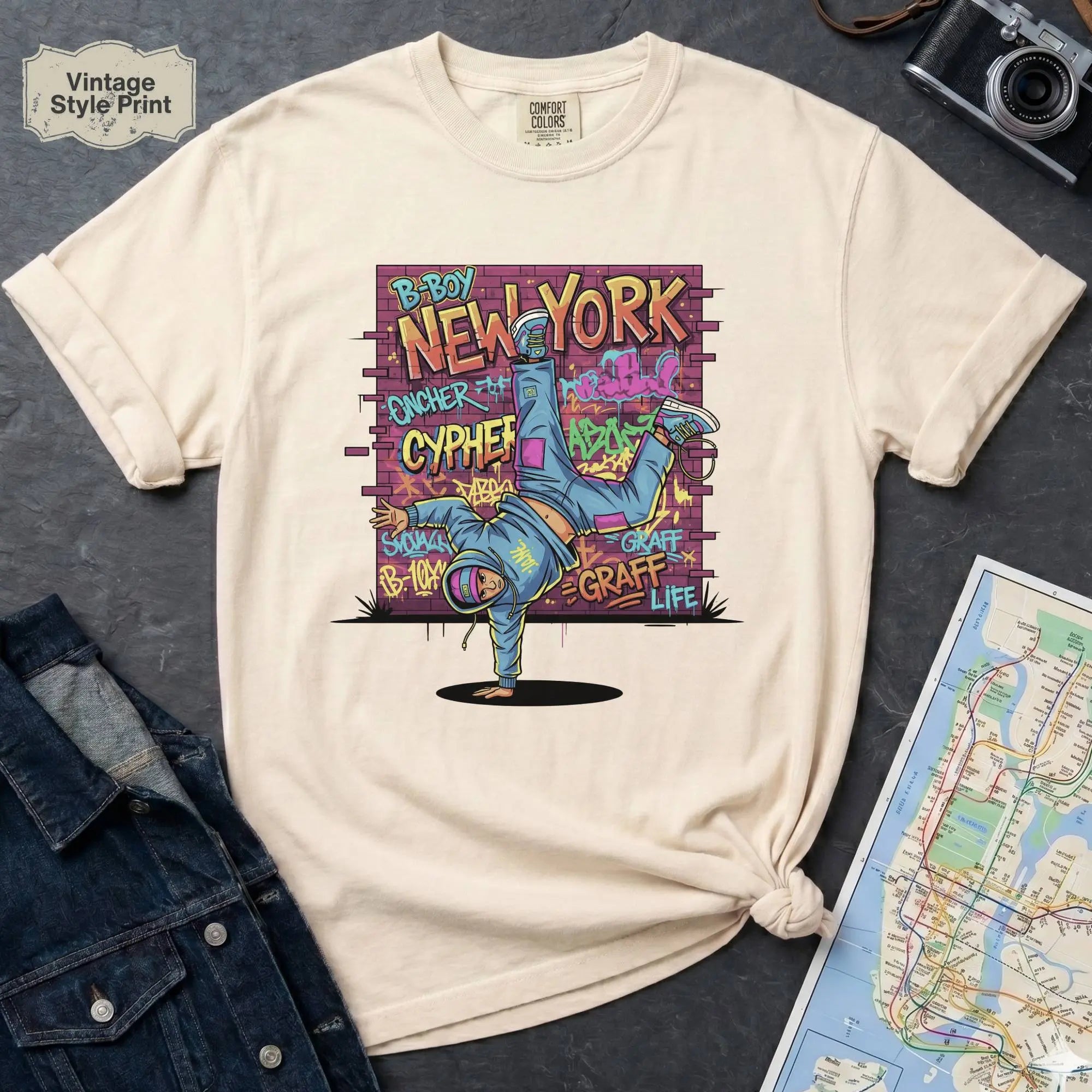 Ivory t-shirt with a colorful graphic design featuring a breakdancer and text on a dark surface with a denim jacket and a map.