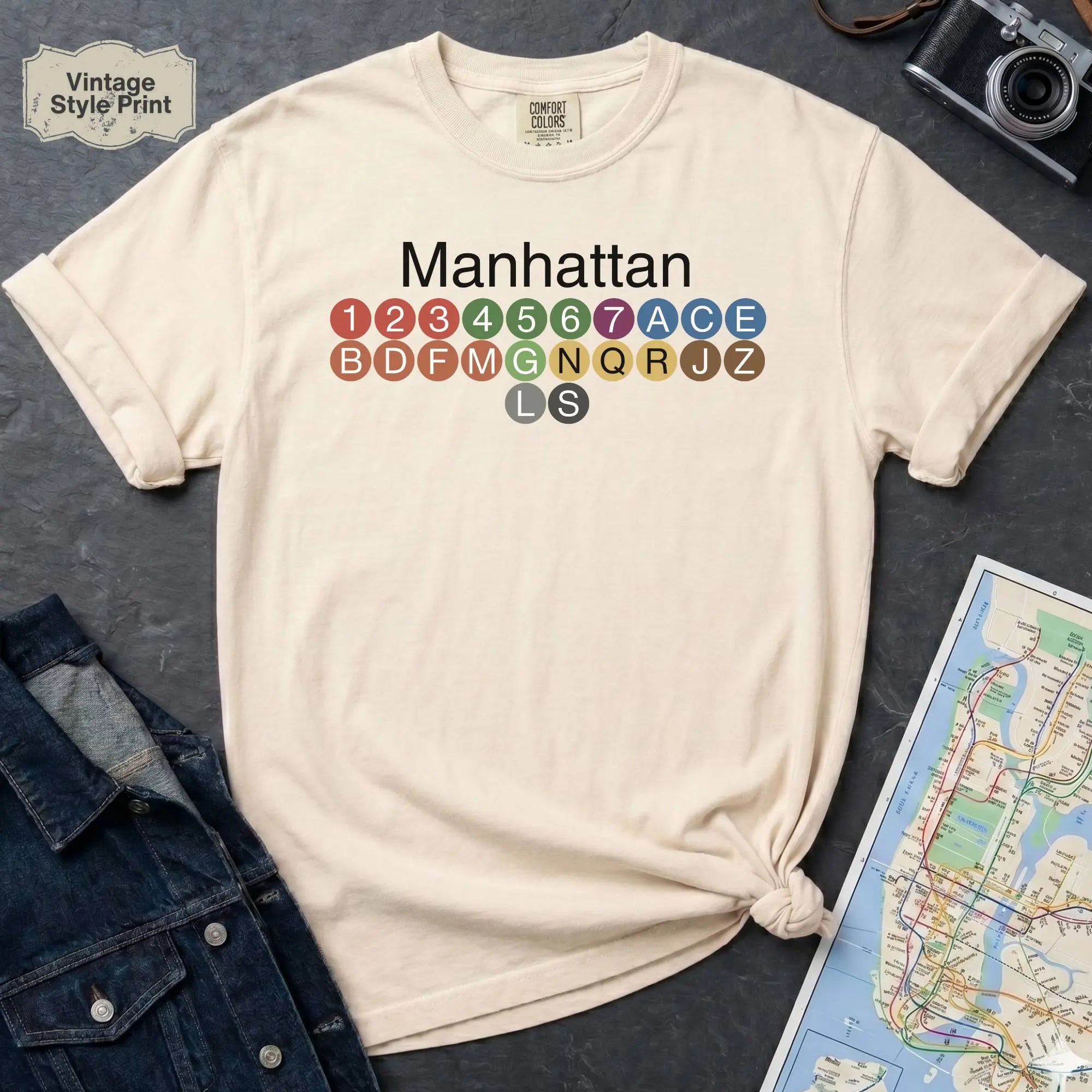 Ivory Comfort Colors t-shirt featuring a minimalist Manhattan subway line graphic with colorful NYC train icons.